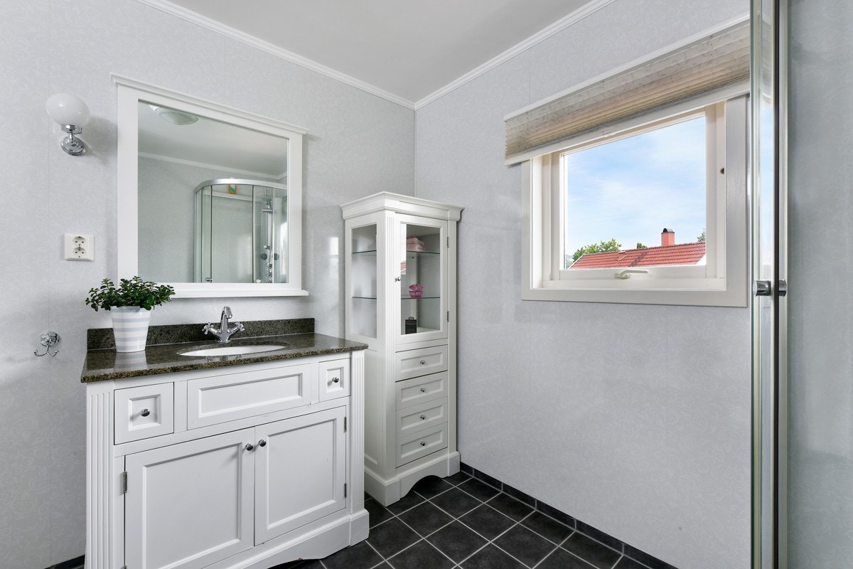 Badet har grå gulv- og sokkelfliser, lyse våtromsplater på veggene, gulvvarme og mekanisk avtrekk. Et vindu gir innslipp av naturlig lys. Galleribilde