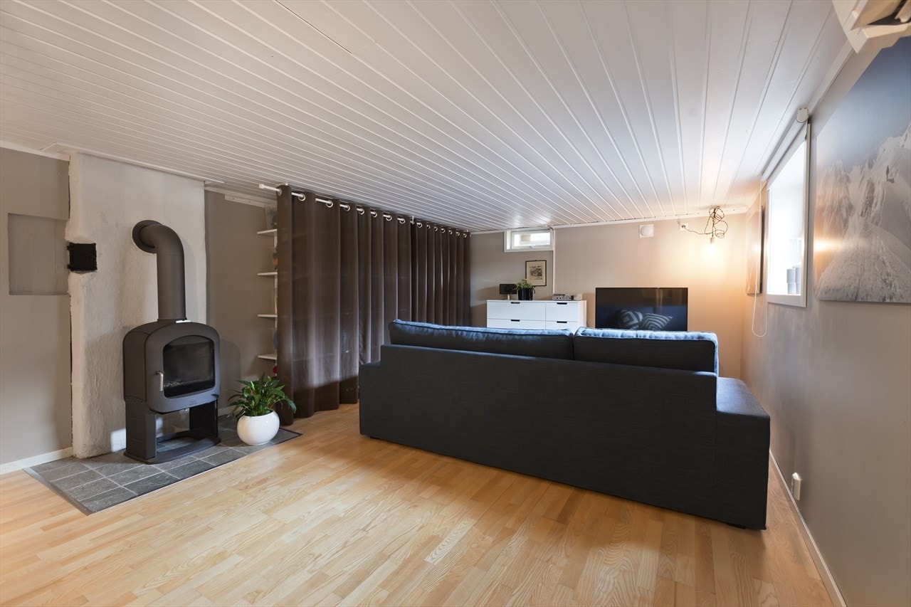 Kjellerstue med god plass til sofa, bord og tv benk. Smart hylleløsning bak gardinene for ekstra lagringsplass. Peisovn for kalde vinterkvelder. Galleribilde