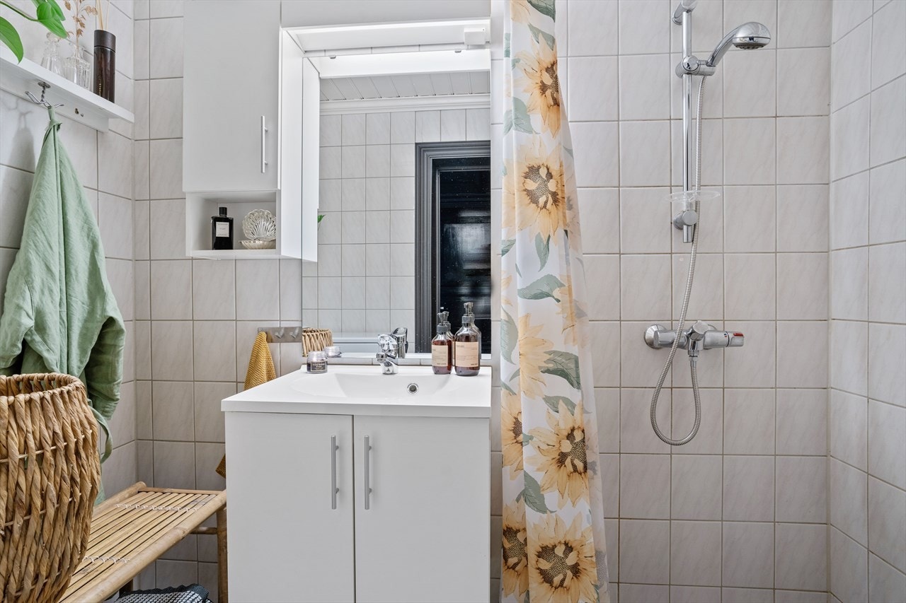 Dusjhjørne avskjermet av forheng. Videre er det helstøpt servant på hvitt underskap med tilhørende speil og veggskap med belysning. Galleribilde