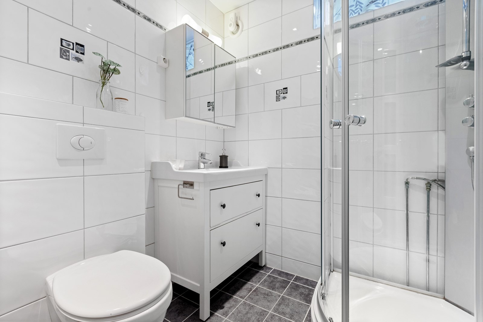 Badet har grå gulvfliser og hvite fliser på veggene, med dekorbård. Galleribilde