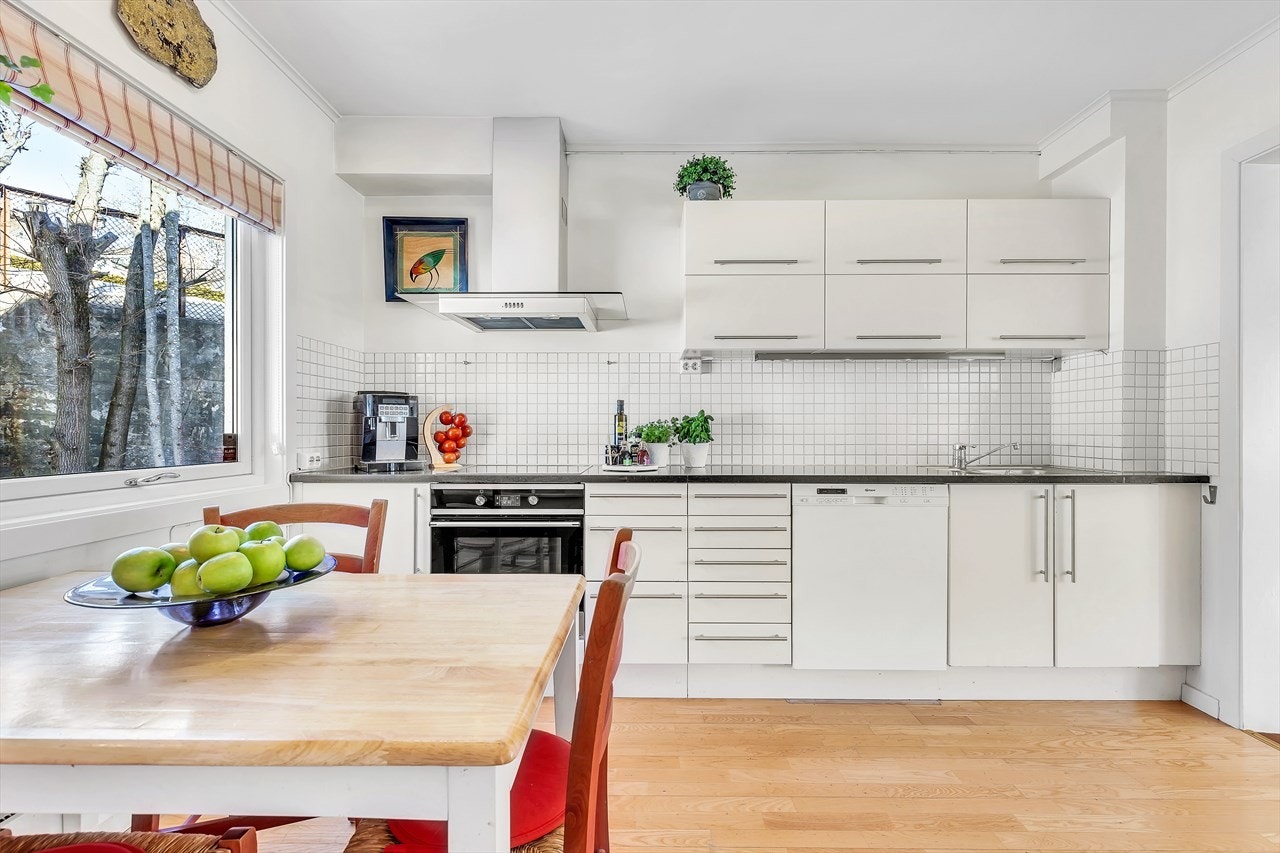 Et romslig og lyst kjøkken med spiseplass ved vindu. Pen, hvit mattlakkert kjøkkeninnredning fra Norema med glatte skapdører og skuffefronter. God med skap- og benkeplass. Avtrekksvifte med kanal ut. Galleribilde