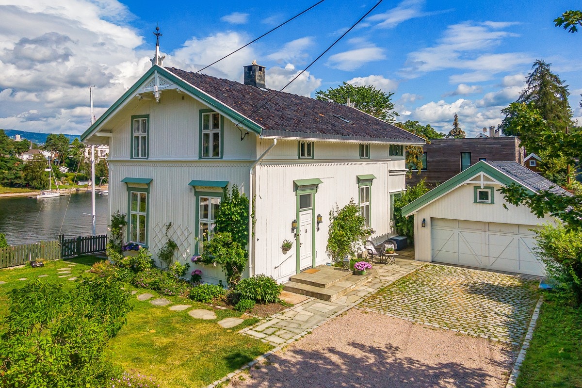 Høyreiste hekker skjermer mot innsyn og ved inngangspartiet klatrer rosebuskene opp etter veggen. Gårdsplassen er gruset, det er brostein i front av garasjen og skiferlagt uteplass ved hovedinngangen. Galleribilde