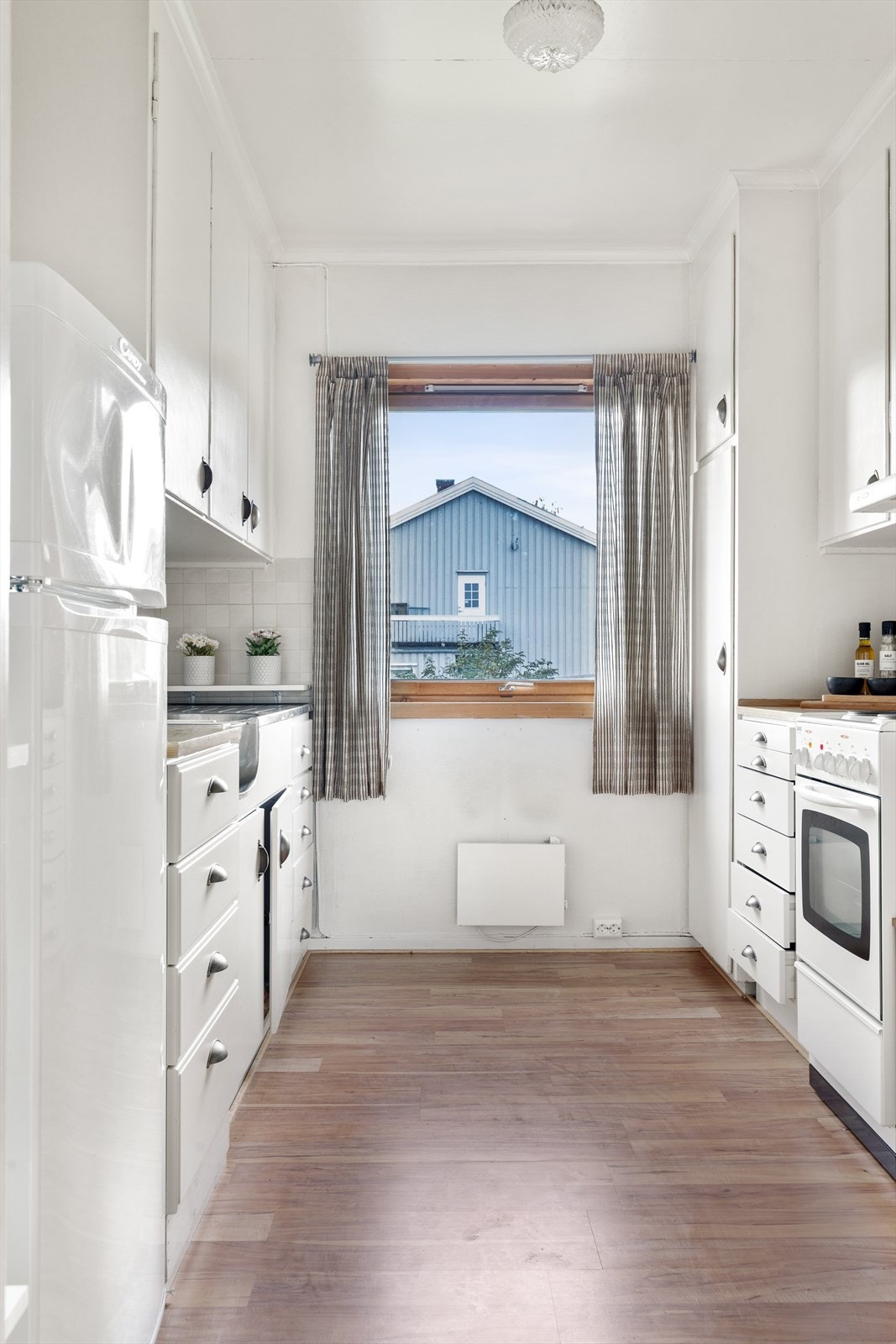 Eldre kjøkkeninnredning med malte skrog og fronter, laminert benkeplate, takhøye overskap, frittstående kjøl- og fryseskap, frittstående mikrobølgeovn og frittstående komfyr. Galleribilde