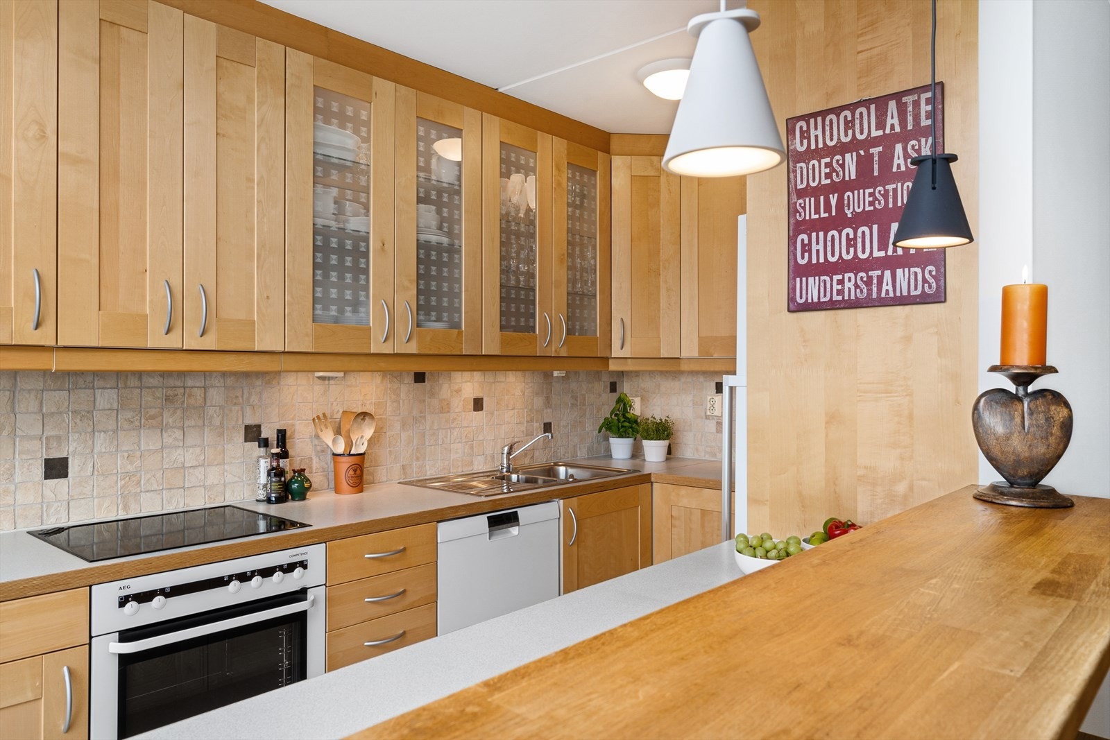 Romslig kjøkken med bjørkefronter. Fliser mellom over- og underskap, og benkeplate med nedfelt vaskekum og keramisk platetopp. Galleribilde