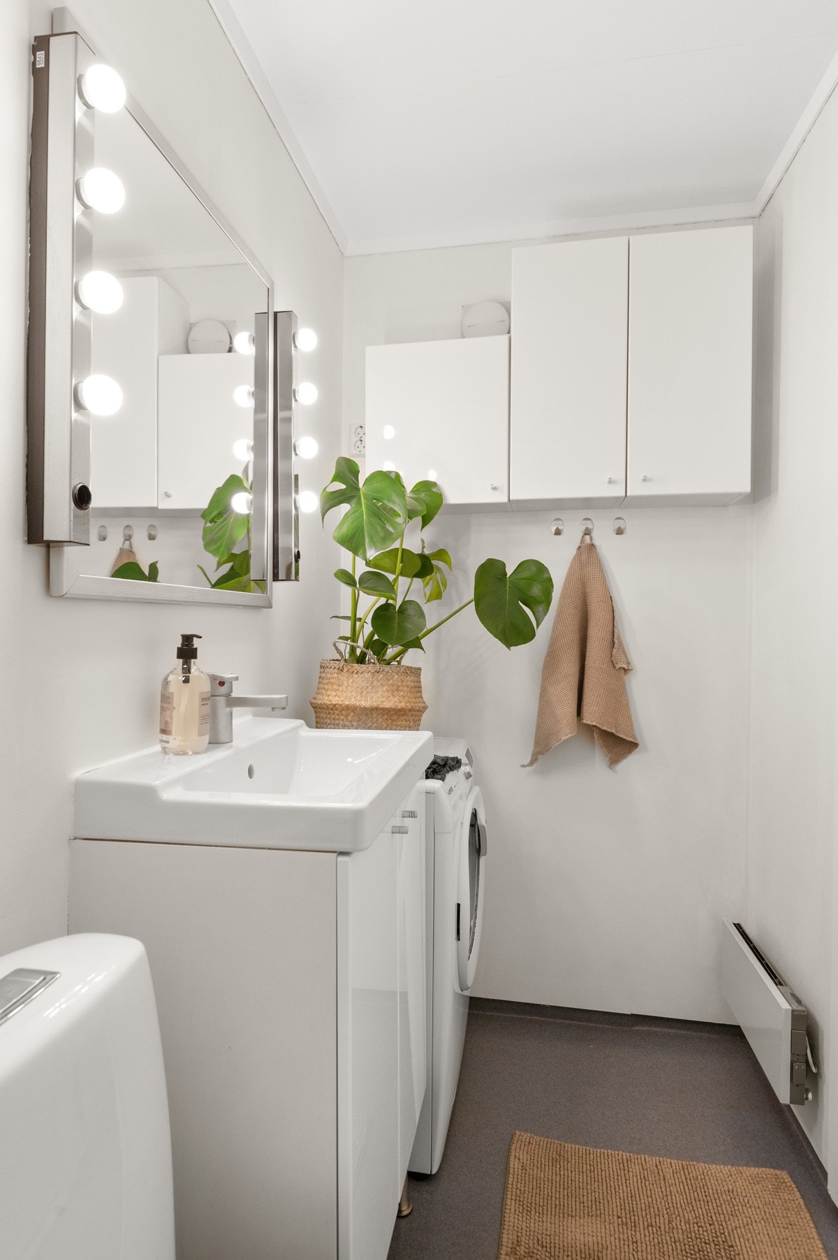 Baderomsinnredning med hvite fronter og heldekkende servant, speil med belysning over, samt ytterligere oppbevaring i vegghengte skap. Galleribilde