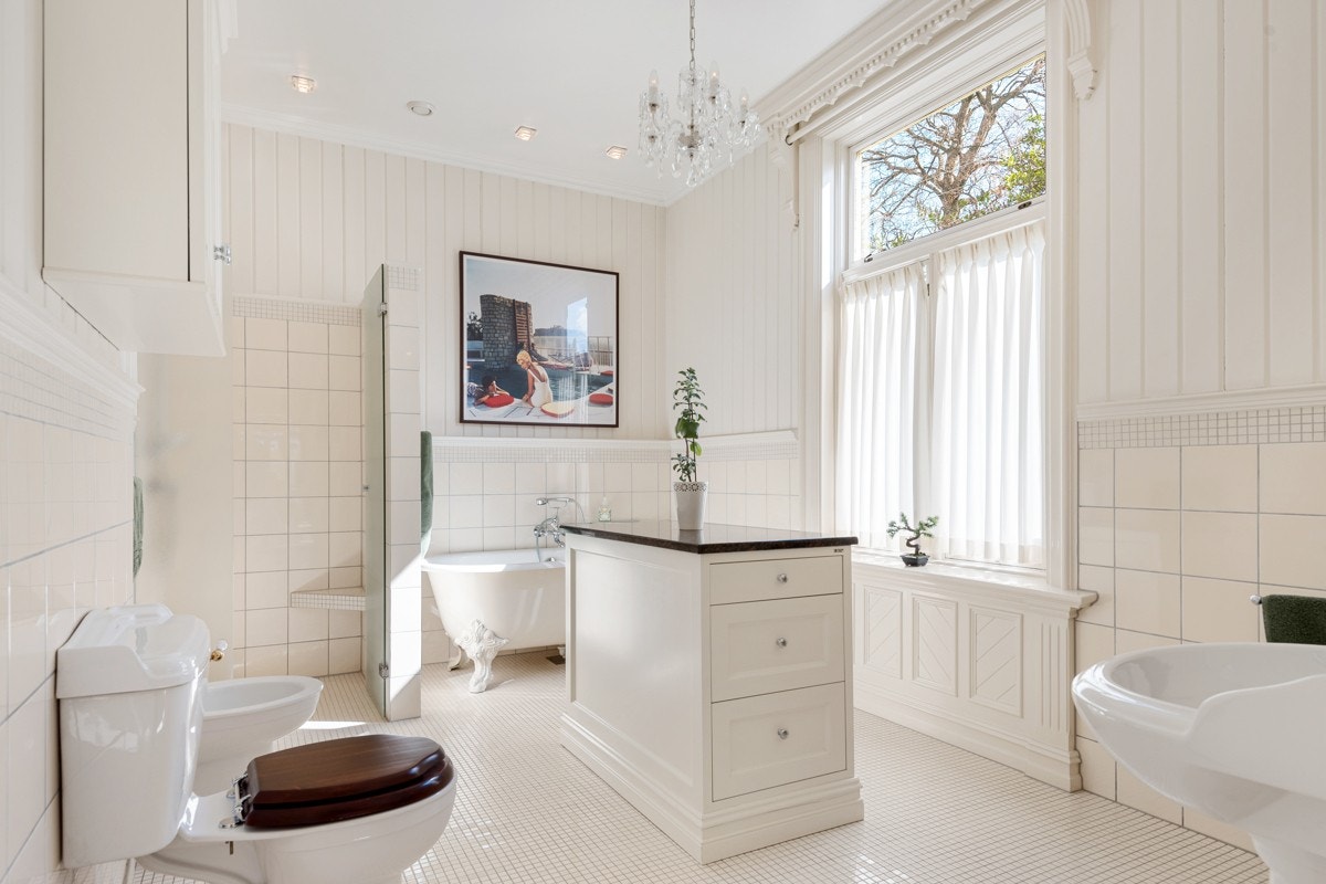 Midt i rommet har et benkeskap fra møbelverksted fått plass. Videre innover er det badekar med løveføtter og en dusjnisje skjermet av glassdør. Badet har også bidét, klosett og to søyleservanter med overhengt speil og belysning. Galleribilde