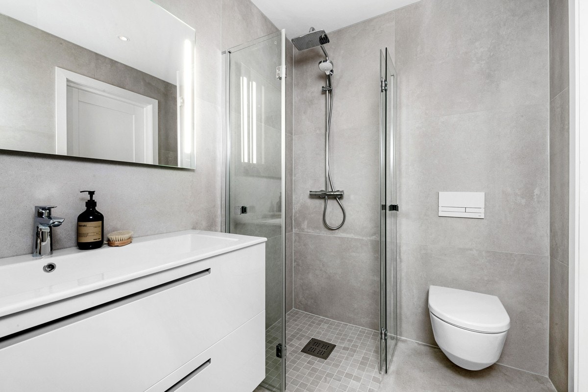 Badene har storformat fliser på gulv og vegger i 90 x 90 cm, varme i gulvet, downlights i taket og vegghengt toalett Galleribilde