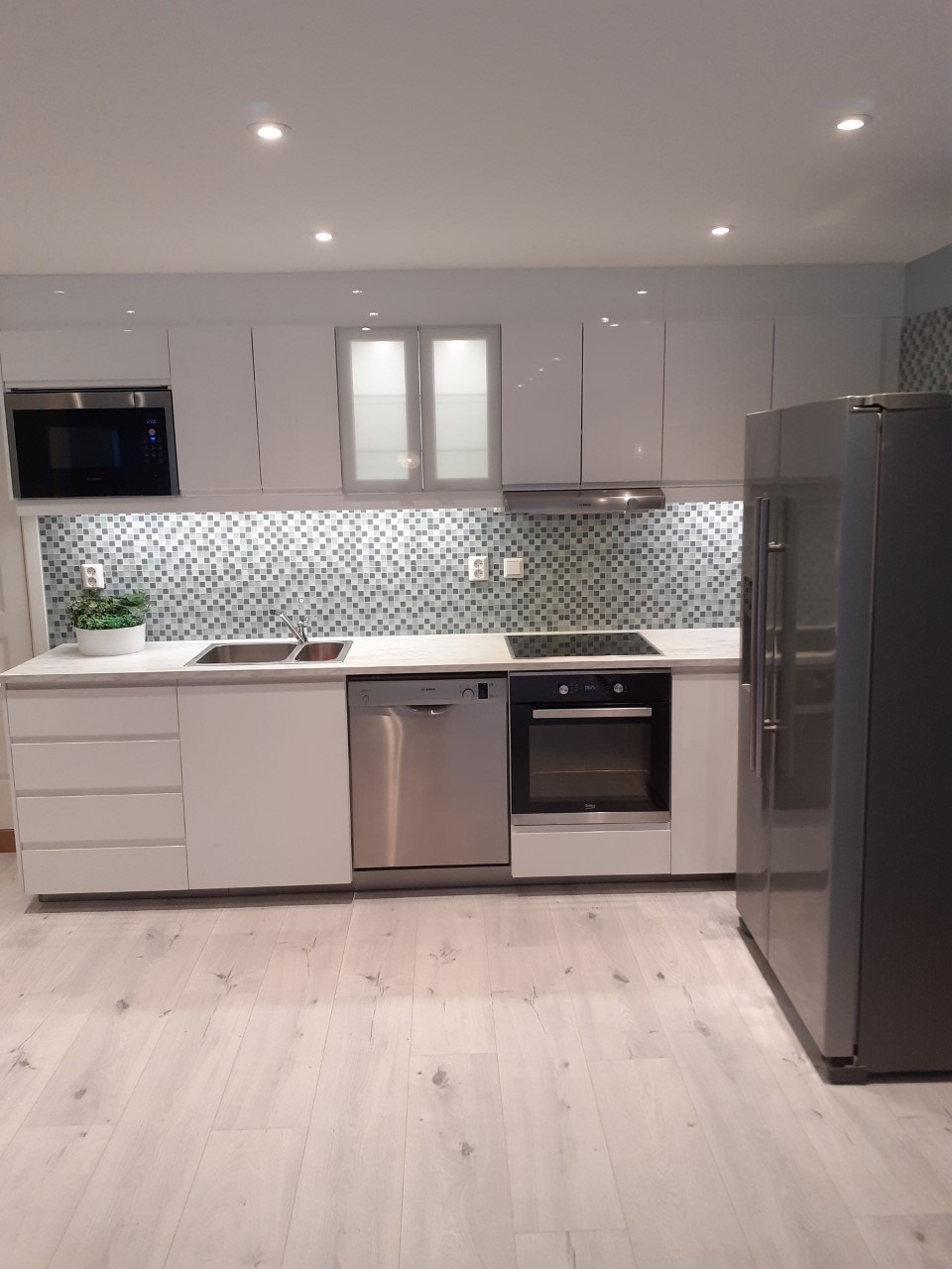 Praktisk kjøkken . Lys i overskap og glassmosaikk fliser på vegg. IKEA kjøkken med høyglans skap og lys over arbeidsbenk. Marmor utseende på benkeplate. Galleribilde