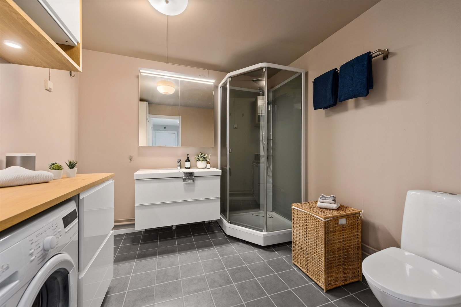 Baderommet er innredet med malte vegger og fliser på gulv. Her er det dusjkabinett, toalett, servant med skuffeseksjon, skap og arbeidsbenk i eik. Under benkeplaten er det skap og opplegg for vaskemaskin. Galleribilde