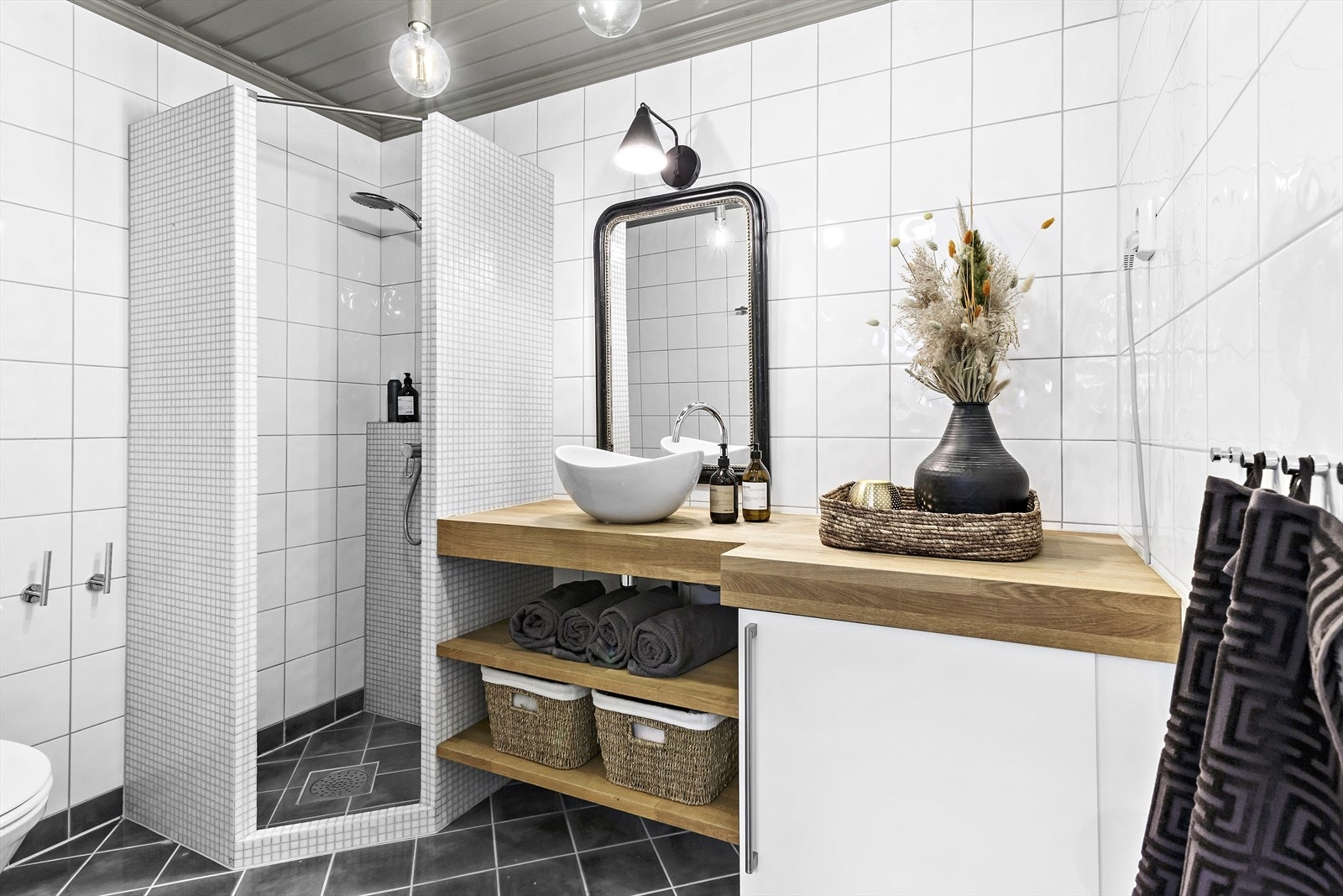 Pent flislagt bad i u.etasje. Innredningen består av servantskap, benkeplate og hyller i tre. Overnpåliggende servant, dusjhjørne og gulvstående toalett Galleribilde