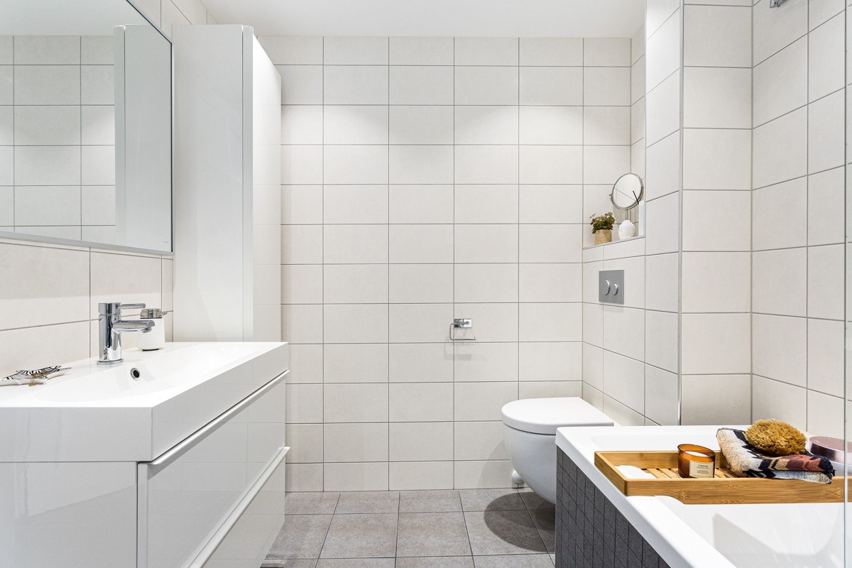 Badet har speil, heldekkende servant på skuffemøblement, vegghengt toalett og innfliset badekar med dusjgarnityr. Galleribilde