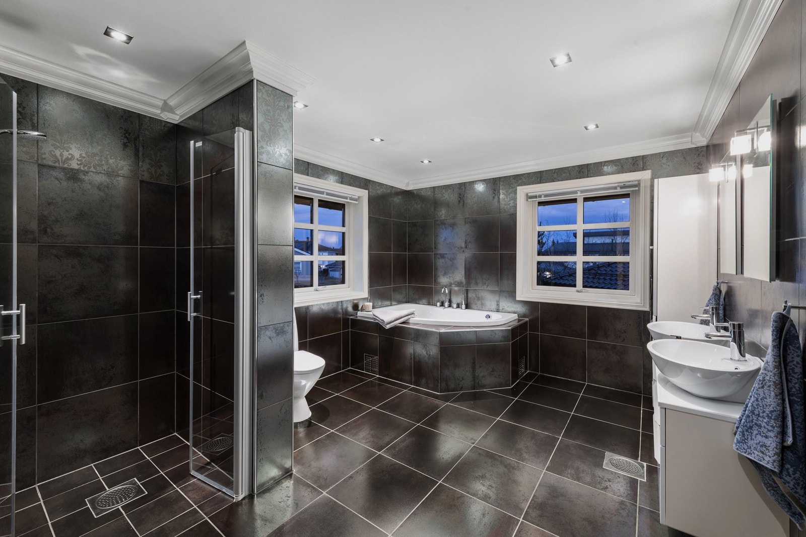 Flott bad med dramatiske, mørke fliser på overflater, med svakt brokademønster på veggene, og vindu på to sider som gir godt med lys Galleribilde