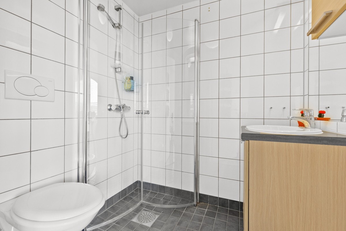 Badet i hybeldelen har fliser på gulv og vegger og varmekabler. Veggmontert toalett, servant med underskap, speil med lys over og dusjhjørne med innfellbare dører. Galleribilde