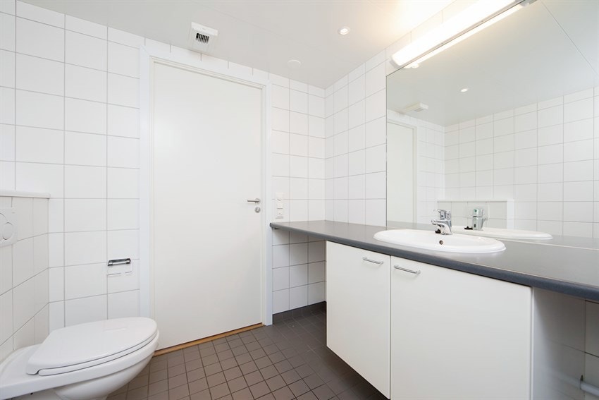 Baderomsinnredningen består av lyse underskap, grå laminatbenkeplate og nedfelt servant. Over benken er det speil og belysning, og under benken er det opplegg og plass for vaskemaskin og tørketrommel. Galleribilde