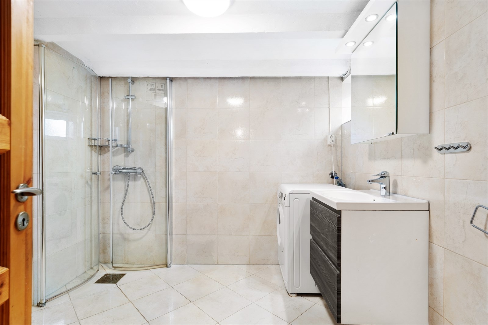 Flislagt baderom som består av toalett, dusj med dusjdører i glass, VVB, opplegg for vaskemaskin og servantinnredning med skuffer under Galleribilde