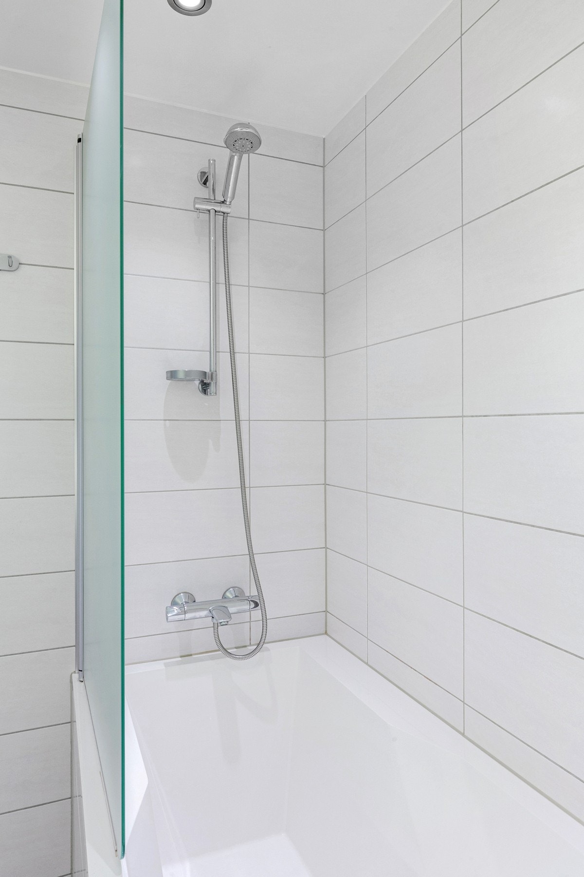 Badet er utstyrt med veggmontert toalett, innfliset badekar, dusjvegg, og veggmontert dusjgarnityr. Galleribilde