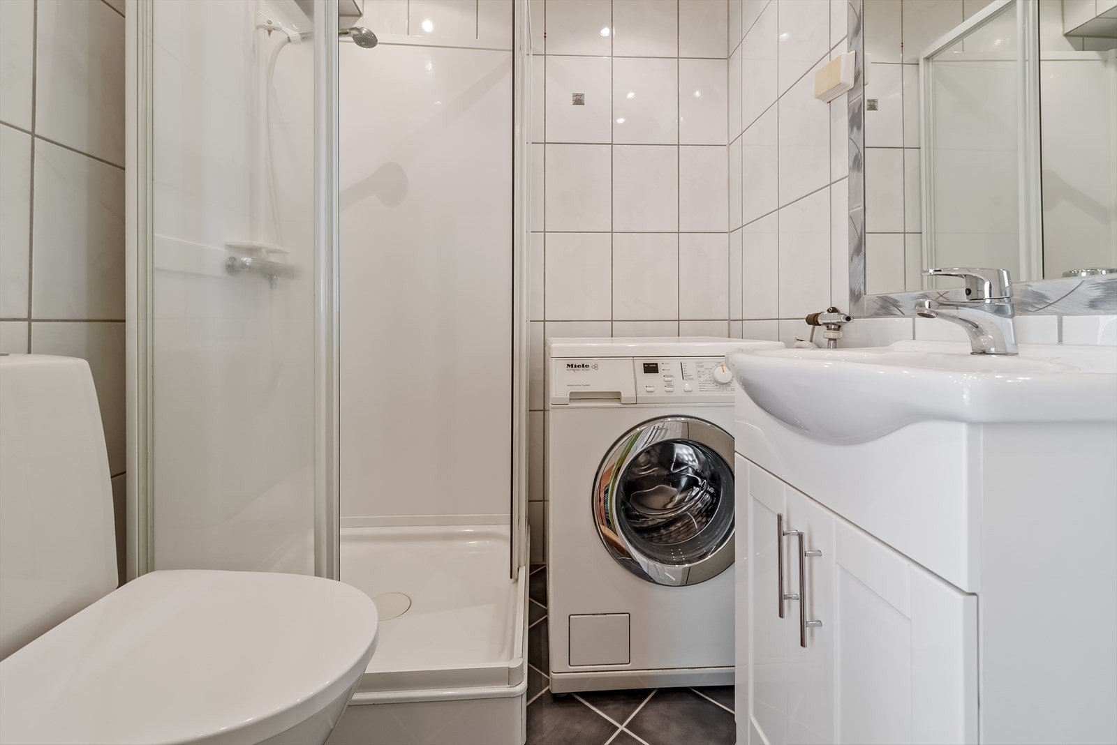 Badet inneholder dusjkabinett fra 2008, wc fra 2018, servantskap med ett-greps blandebatteri, innmurt speil på vegg, belysning i himling med innfelte spotter og opplegg for vaskemaskin. Galleribilde