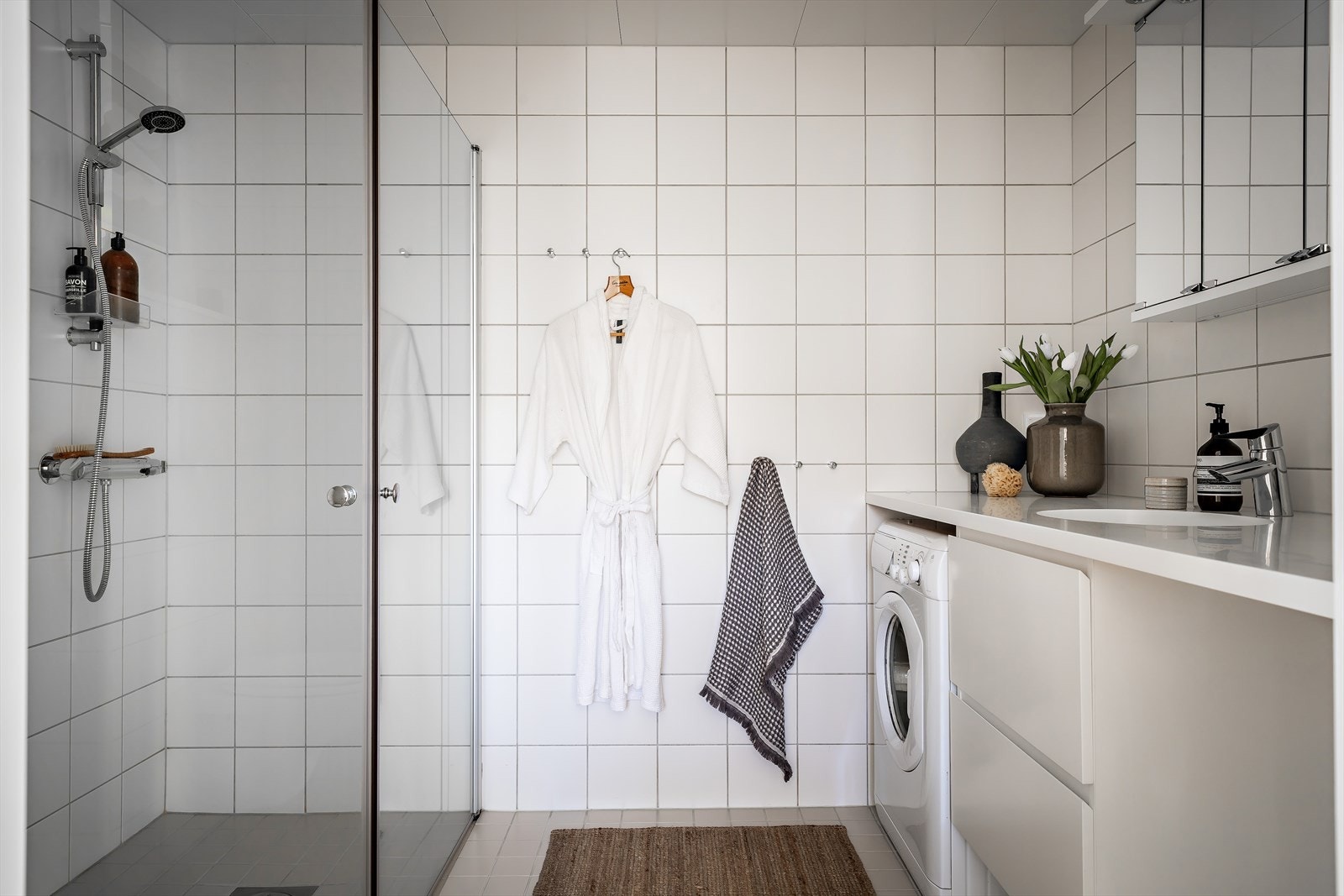 Baderommet har et pent, stilrent uttrykk med hvite veggfliser og lysegrå gulvfliser med gulvvarme. Himling er pent lyssatt med downlights. Under benken er det nisje med opplegg for vaskemaskin. Galleribilde