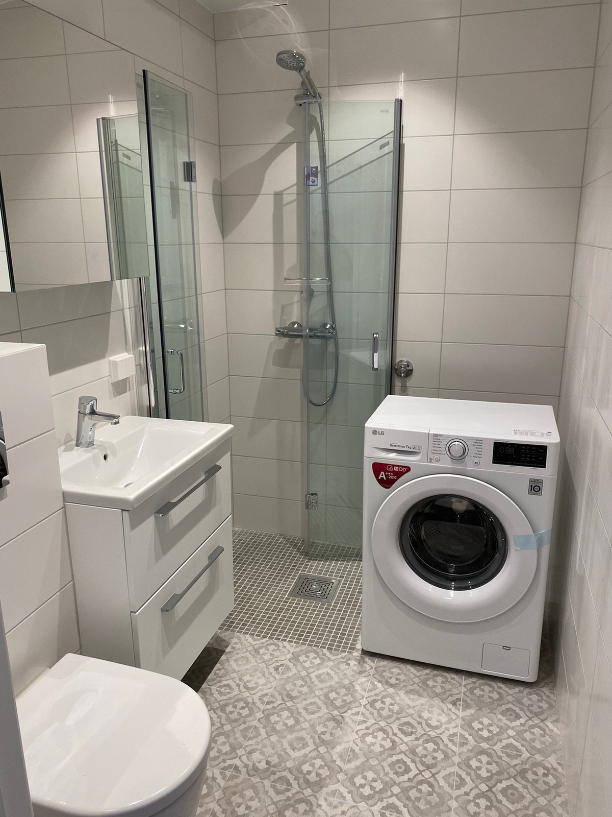 Bad med foldbare dusjdører, vegghengt toalett og vaskemaskin. (bildet er av en annen vaskemaskin, og det er montert en hylle over vaskemaskinen i dag) Galleribilde