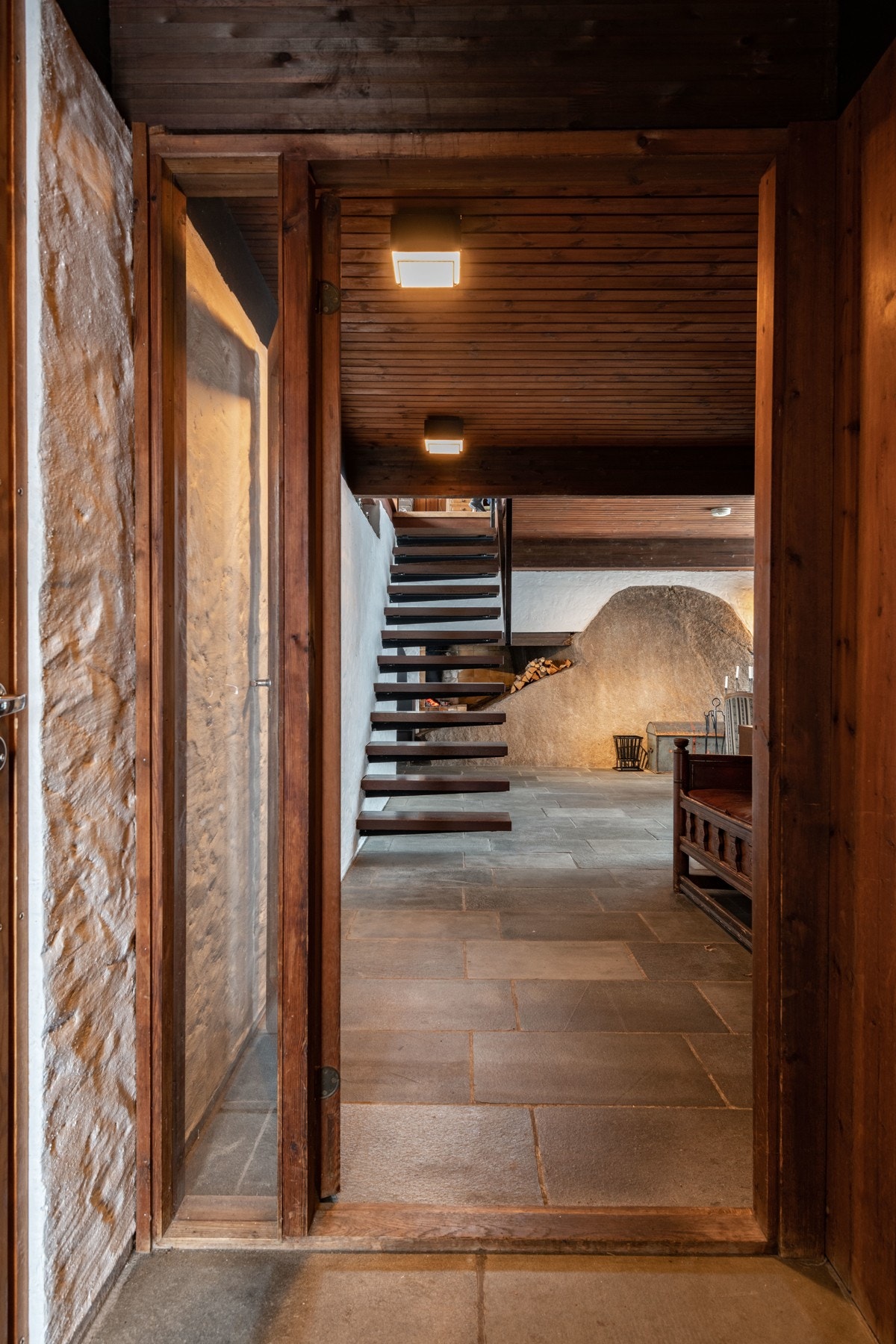 En glassdør stenger vindfanget fra kjellerstuen, men utsikten mot den unike peisen som er integrert i grunnfjellets form i peisestuen blir et blikkfang fra det øyeblikk man åpner ytterdøren. Galleribilde