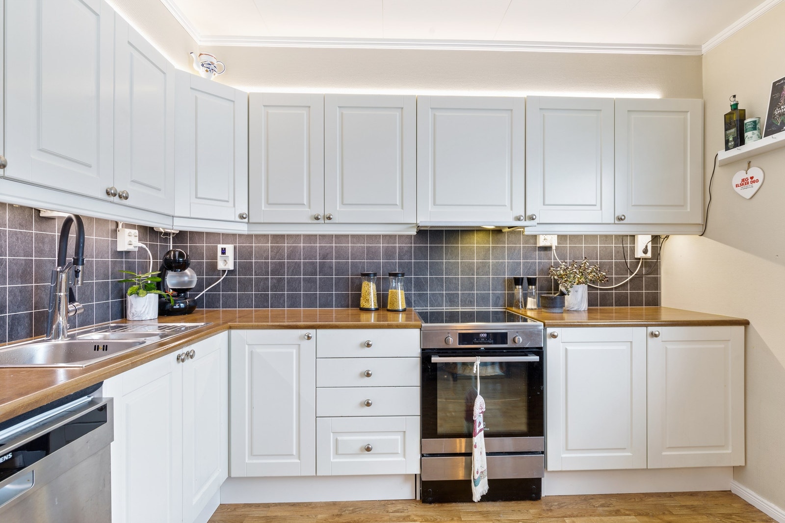 Hvit profilert innredning med laminerte benkeplater. Opplegg for oppvaskmaskin, ventilator og kitchenboardplater mellom overskap/benk.
Blandebatteri med automatisk avstengning til oppvaskmaskin. Galleribilde