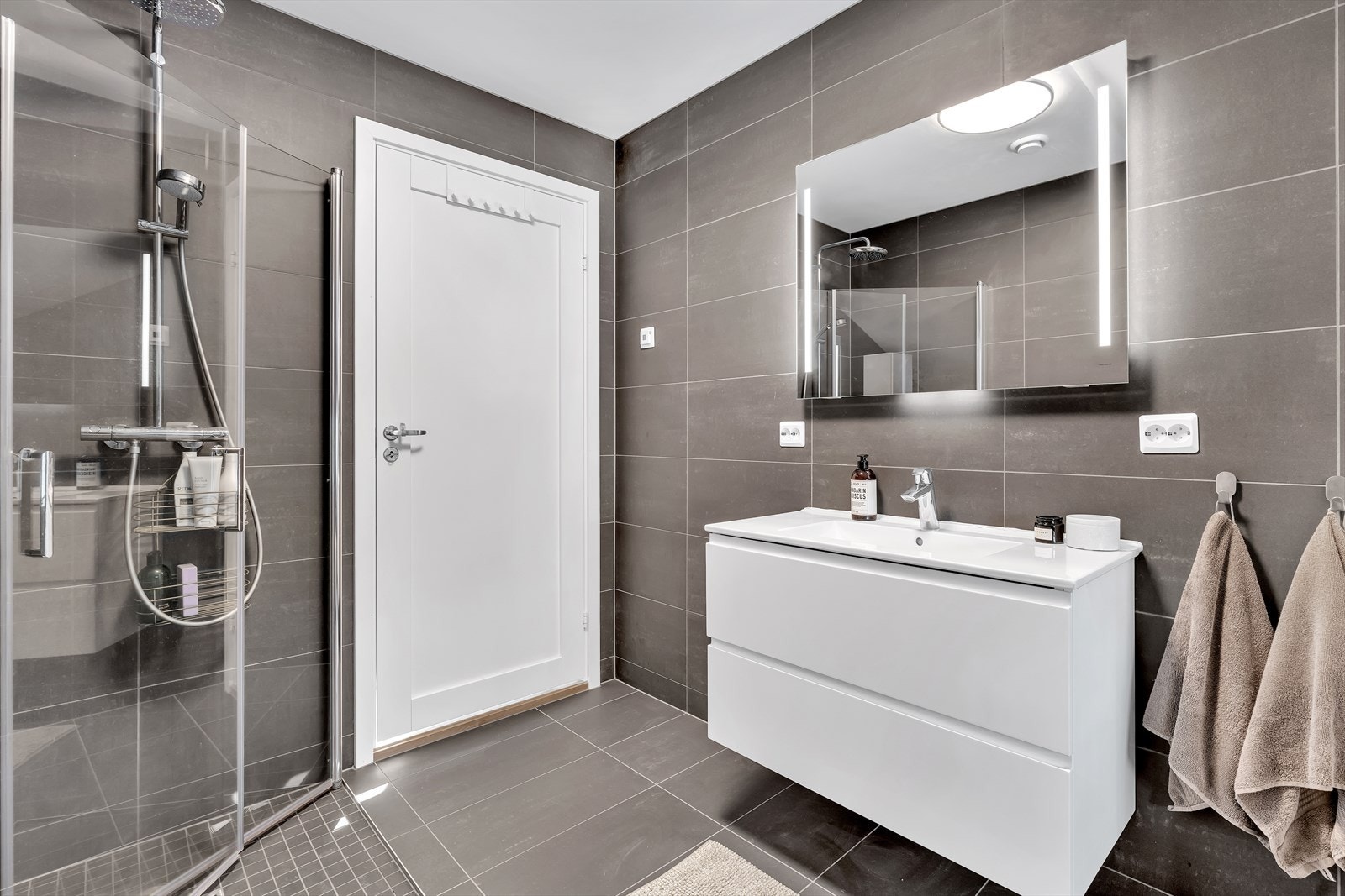 Badet er utstyrt med dusjnisje med innfellbare glassdører, vegghengt wc og servantinnredning med skuffer. Galleribilde