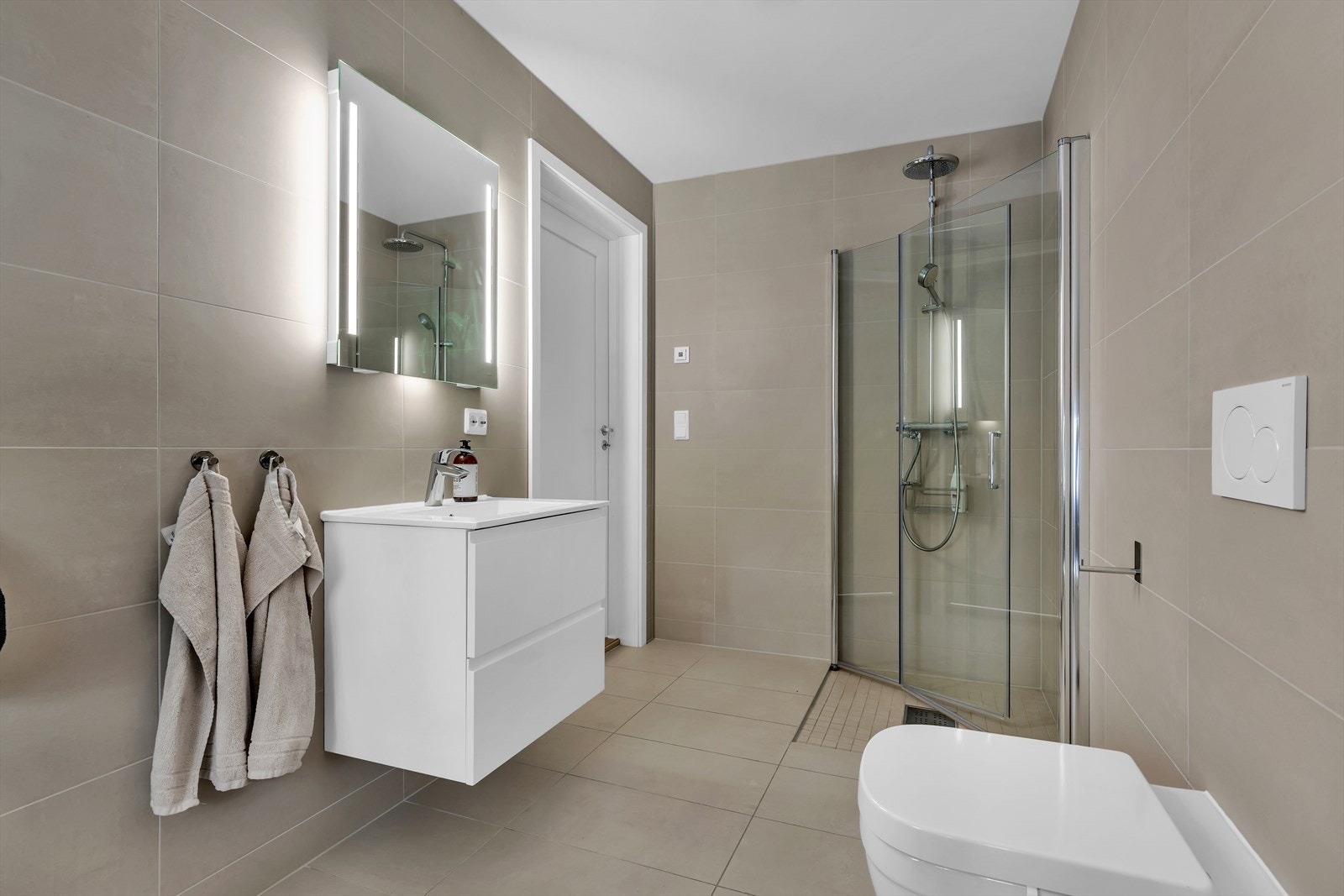 Badet er utstyrt med dusjnisje med innfellbare glassdører, vegghengt wc og servantinnredning med skuffer. Galleribilde