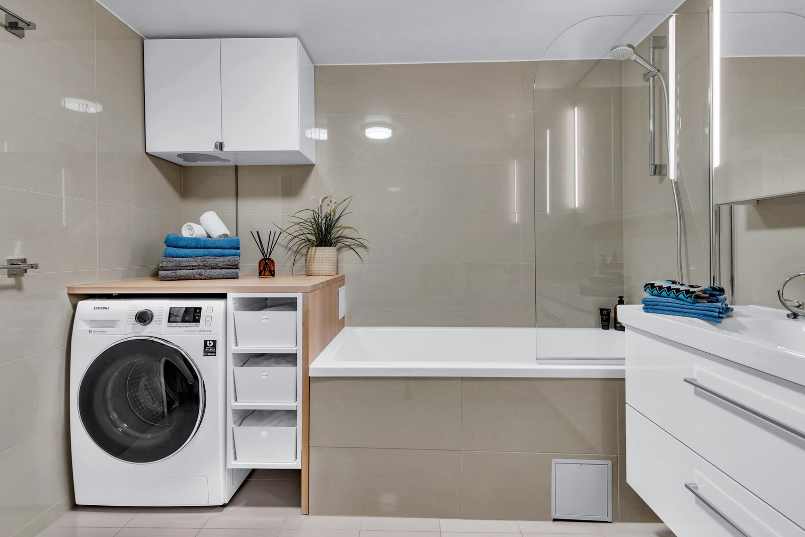 Badet er innredet med dusj i badekar med en hengslet glassdør, servantskap med ett-greps blandebatteri og heldekkende servant, overskap med og uten speil, arbeidsbenk ved vaskemaskin og gulvstående toalett. Galleribilde