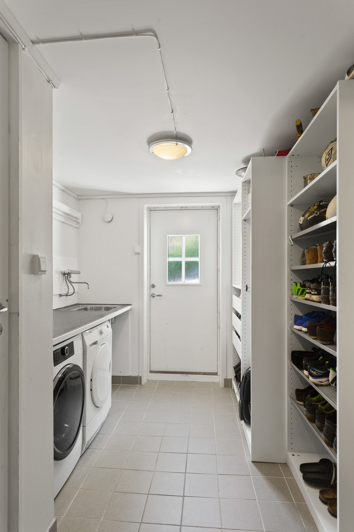 Det praktiske vaskerommet har fliser på gulvet og ny benkeplate, som er plassert over vaskemaskin og tørketrommel. Det er rikelig med hylleplass og heng for lagring av sko og klær. Veggene er malt lyse, og det er dør ut til område nedenfor uteboden. Galleribilde