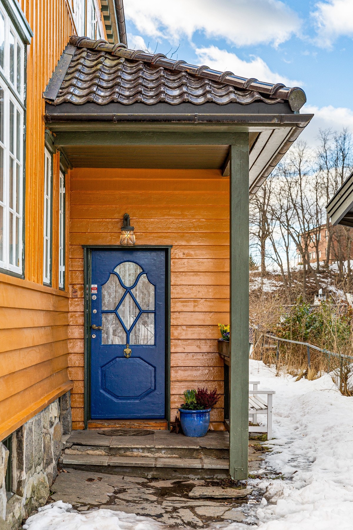 Fra gårdsplassen går det trinn opp til overbygget inngangsparti med belysning. En blåmalt dør fører inn i vindfanget. Galleribilde