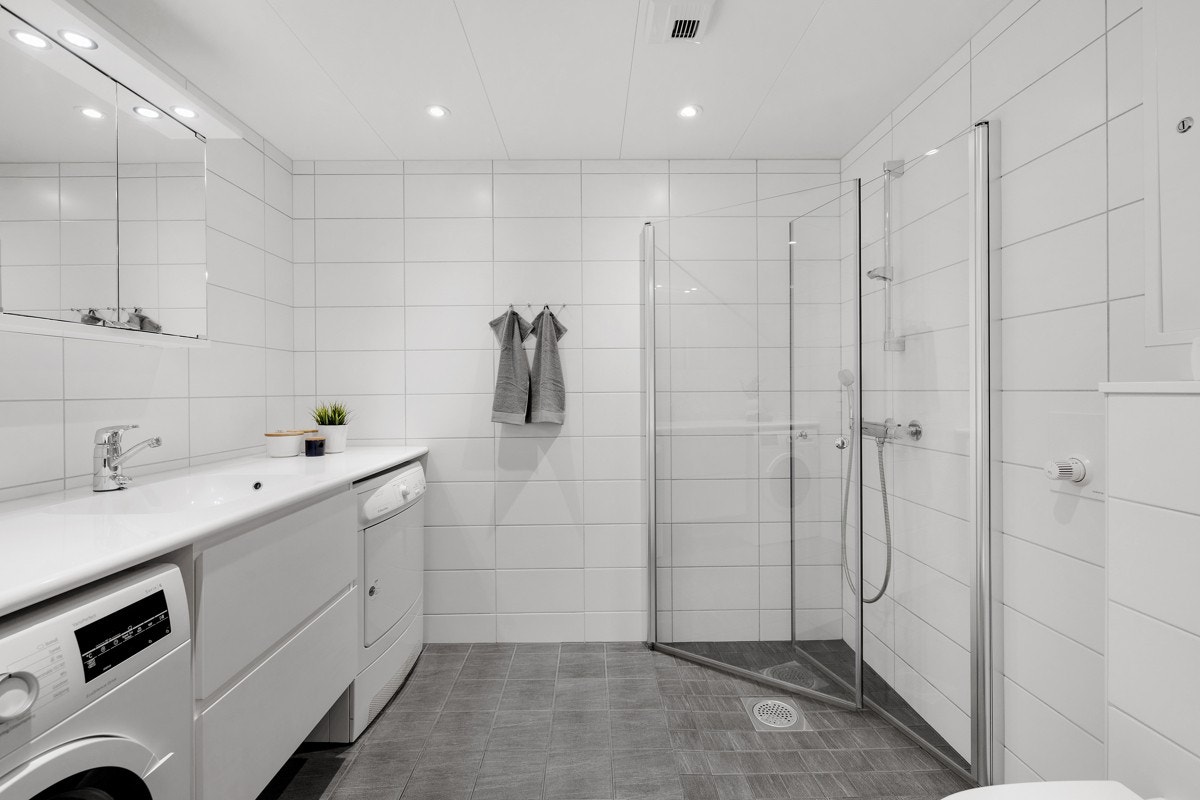 Stort og pent flislagt bad som bl.a. inneholder hvit moderne baderomsinnredning med skuffeløsninger og med speil og lysarmatur, vegghengt toalettskål og dusjnisje med dusjdører i herdet glass Galleribilde