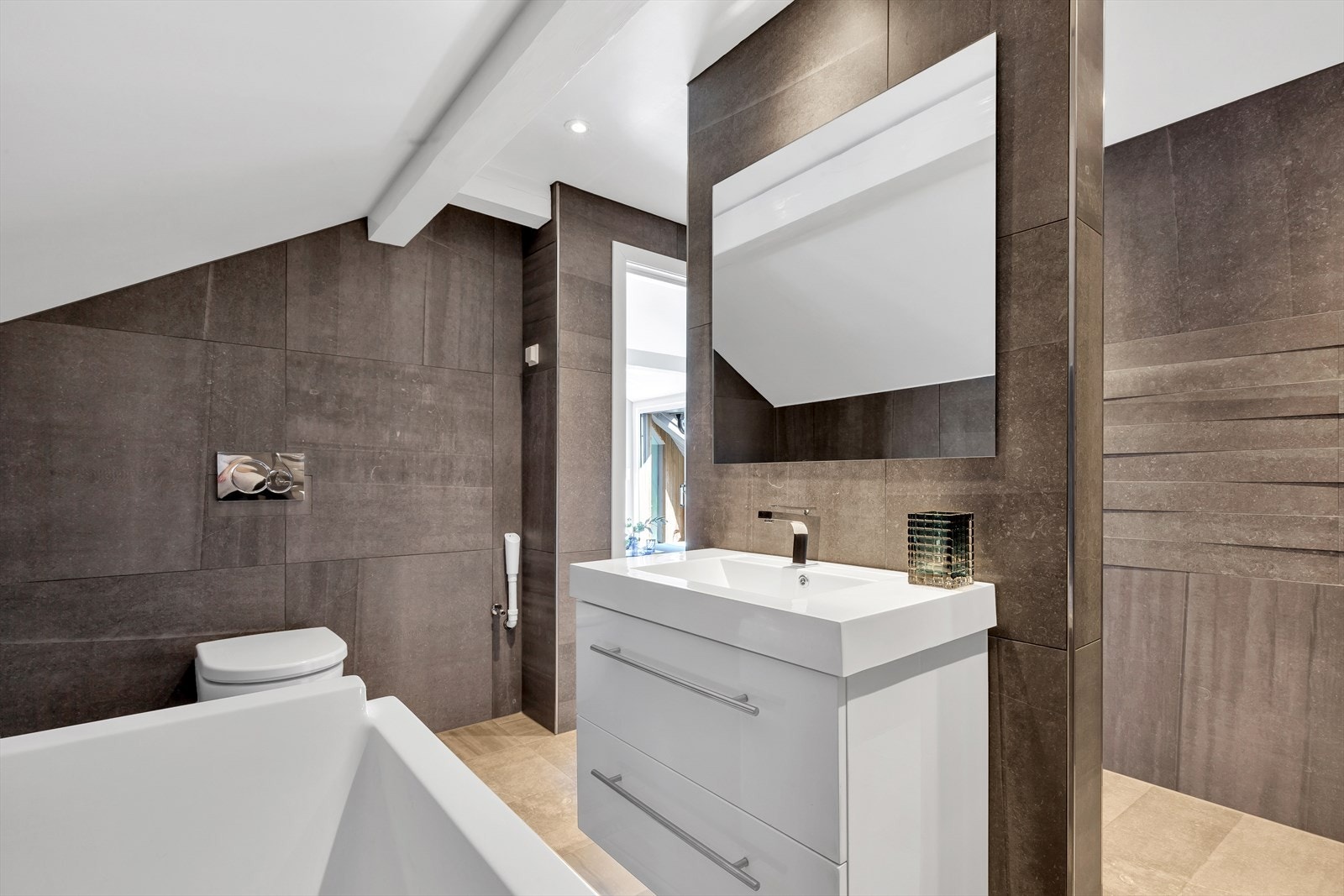 Ellers har badet veggmontert wc og servant over innredning med skuffer. Det er fliser med varme på gulv som sørger for en lun og god temperatur og det er downlights i himling. Galleribilde