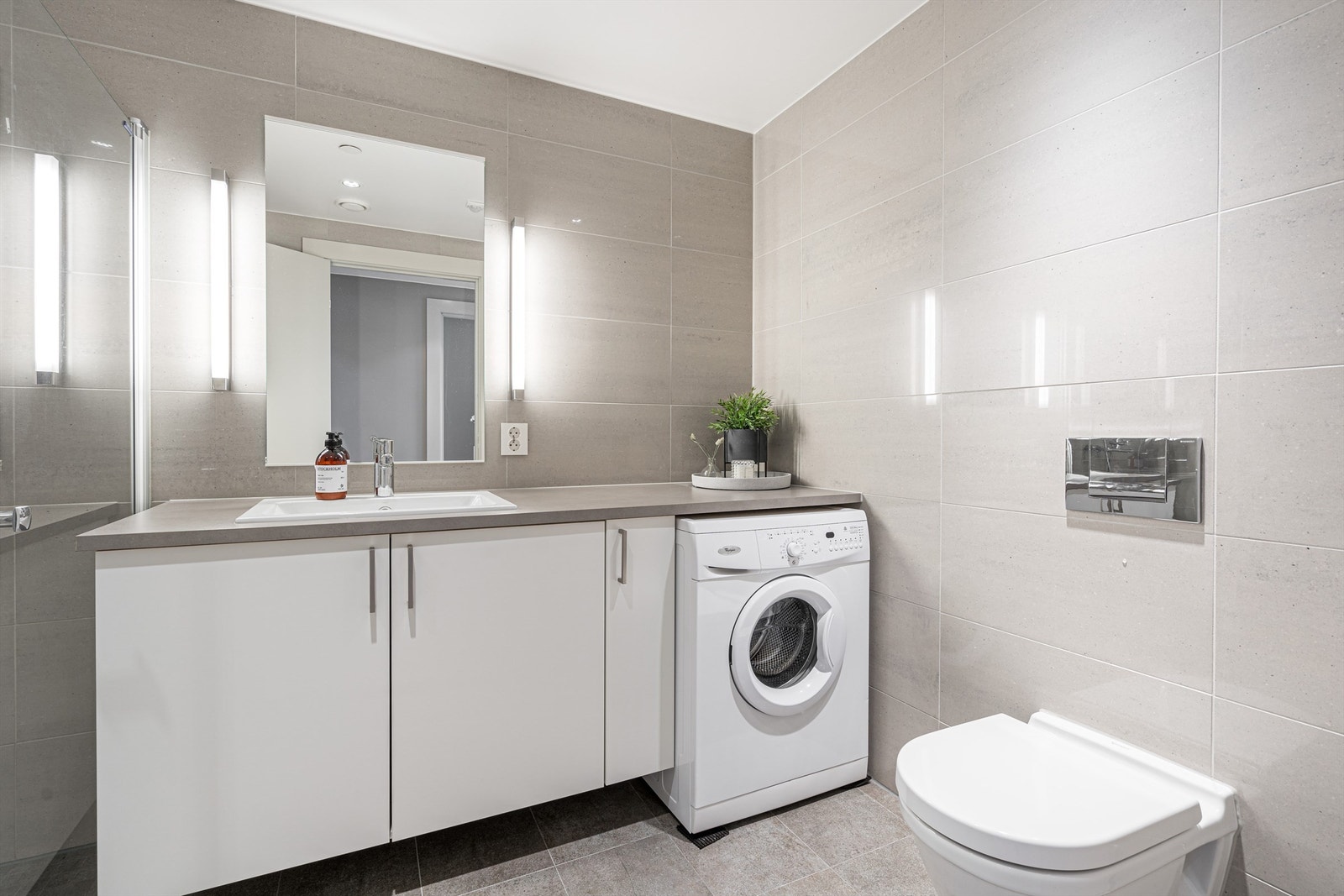 Hovedbadet fremstår lyst og innbydende. Benkeplate med nedfelt servant, skapinnredning, speil og opplegg for vaskemaskin eller kombimaskin. Galleribilde