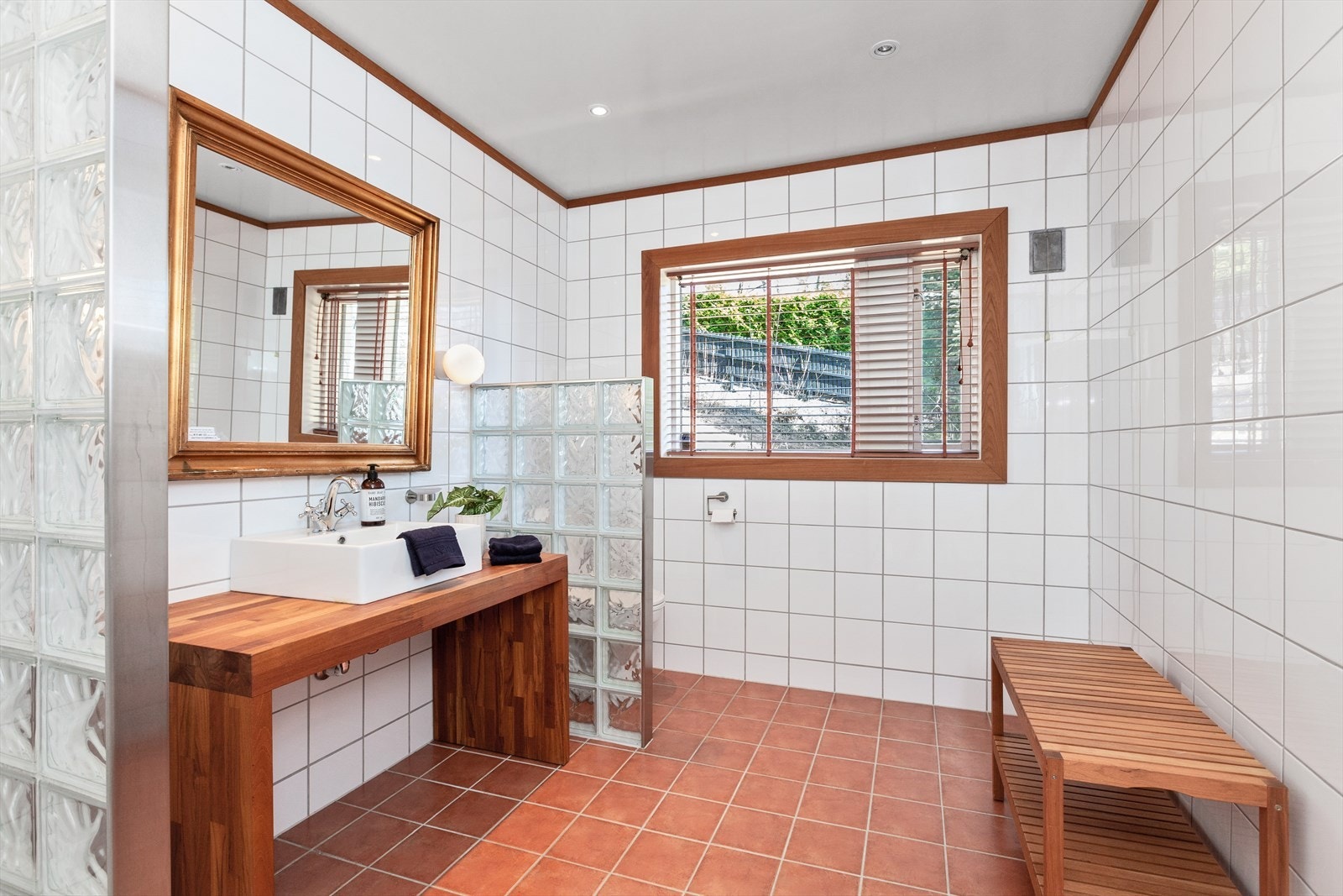 Flislagt bad med varmekabler i gulvet og downlights i taket. Dusjssonen er skjermet med glassvegger av glassbyggerstein. Gulvstående toalett og toppmontert servant. Galleribilde