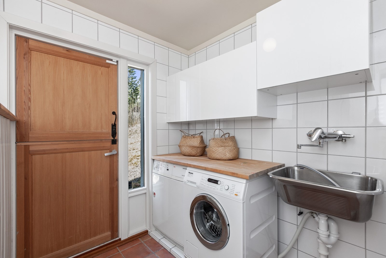 Flislagte gulv og vegger, med hvite, slette overskap. Benkeplate i heltre. Stålkum med ett-greps blandebatteri. Opplegg til vaskemaskin og tørketrommel. Galleribilde