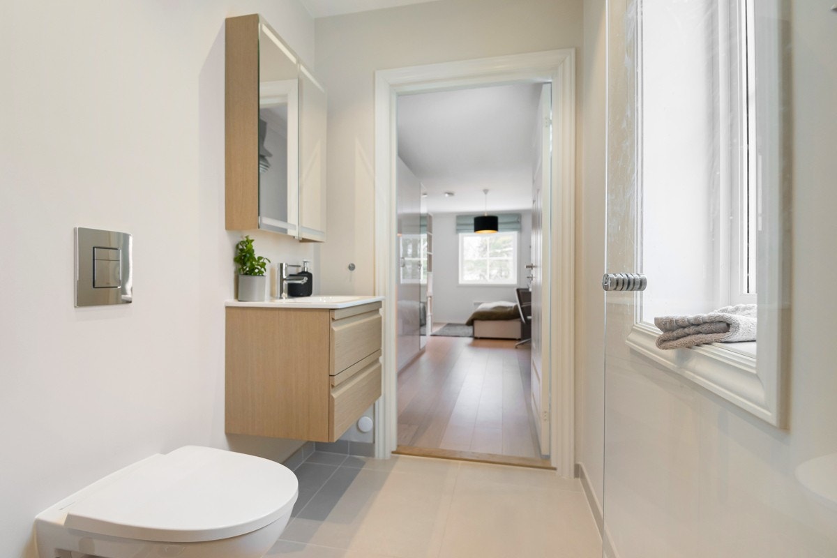 Dette badet har store, sandfargede gulvfliser, sokkelflis og fliser i dusjsonen. Veggene for øvrig er malt i en lys fargevalør. Baderomsmøblementet fra Dansai er i eikefarget utførelse. Det består av heldekkende servant på skuffeseksjon og et speilskap. Galleribilde