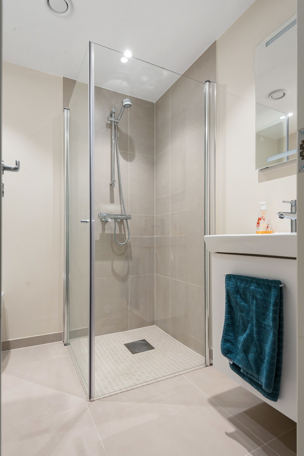 Bad med dusjhjørne, vegghengt toalett, og opplegg for vaskemaskin i plassbesparende vaskesøyle. Baderomsinnredningen er i hvit, glatt utførelse og består av heldekkende servant på skuffeseksjon og speil med integrert belysning. Galleribilde