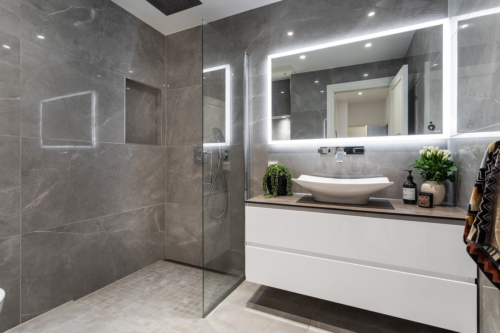 Flislagt baderom fra 2019 med gulvvarme og nedsenket himling med downlights. Vegghengt servantskap i slett utførelse, servant på benkeplate med integrert to-greps armatur fra Hansgrohe og speil med belysning og stikkontakter i vegg over servant Galleribilde