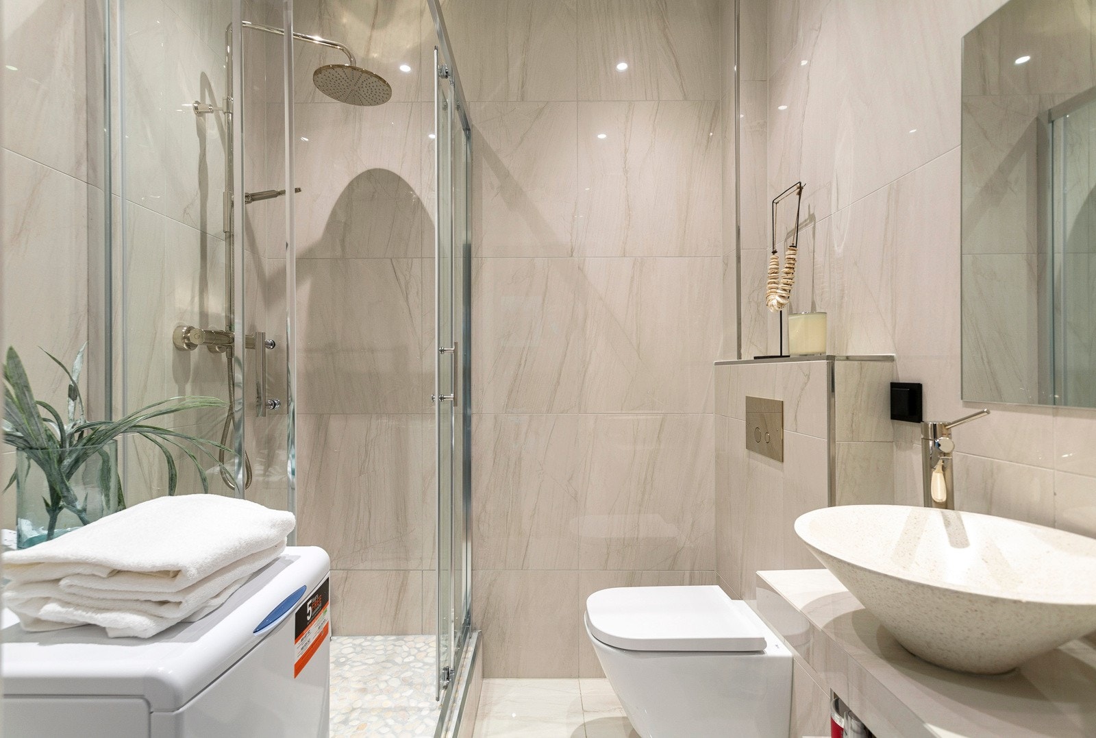 På badet er det vegghengt toalett og opplegg for vaskemaskin. Et oppforet dusjhjørne har regnfallsdusj, rullestein på gulvet og skyvedører i herdet glass. Den øvrige innredningen består av et speil og en flislagt hylleløsning med toppmontert servantskål. Galleribilde