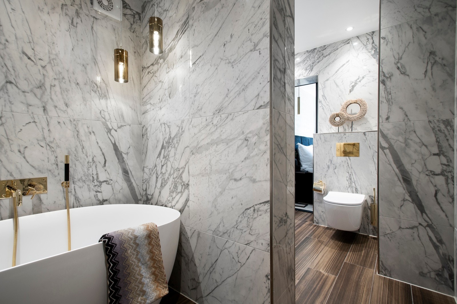 Badet ved soverommet har store, lyse veggfliser med marmorlook og gulvfliser som ligner parkett. Det er gulvvarme, vegghengt toalett og downlights i himlingen. I en egen nisje har badet vakre taklamper, et nedsenket dusjhjørne og et flott badekar. Galleribilde