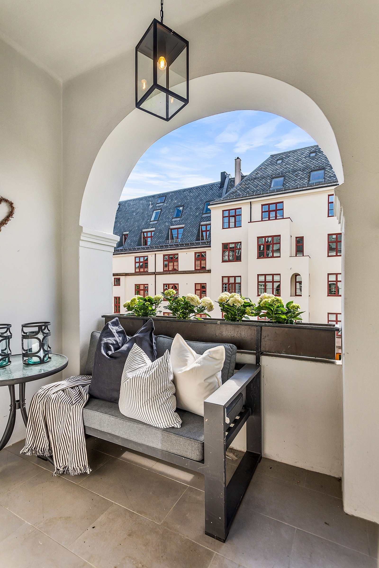 Østvendt, overbygget balkong på ca. 3 kvm med adkomst fra dagligstuen. Balkongen har grå gulvfliser i 30 cm x 60 cm, stilfull bueåpning mot gaten, og rekkverk i pusset og malt mur. Det er montert blomsterkasse på toppen av rekkverket. Galleribilde