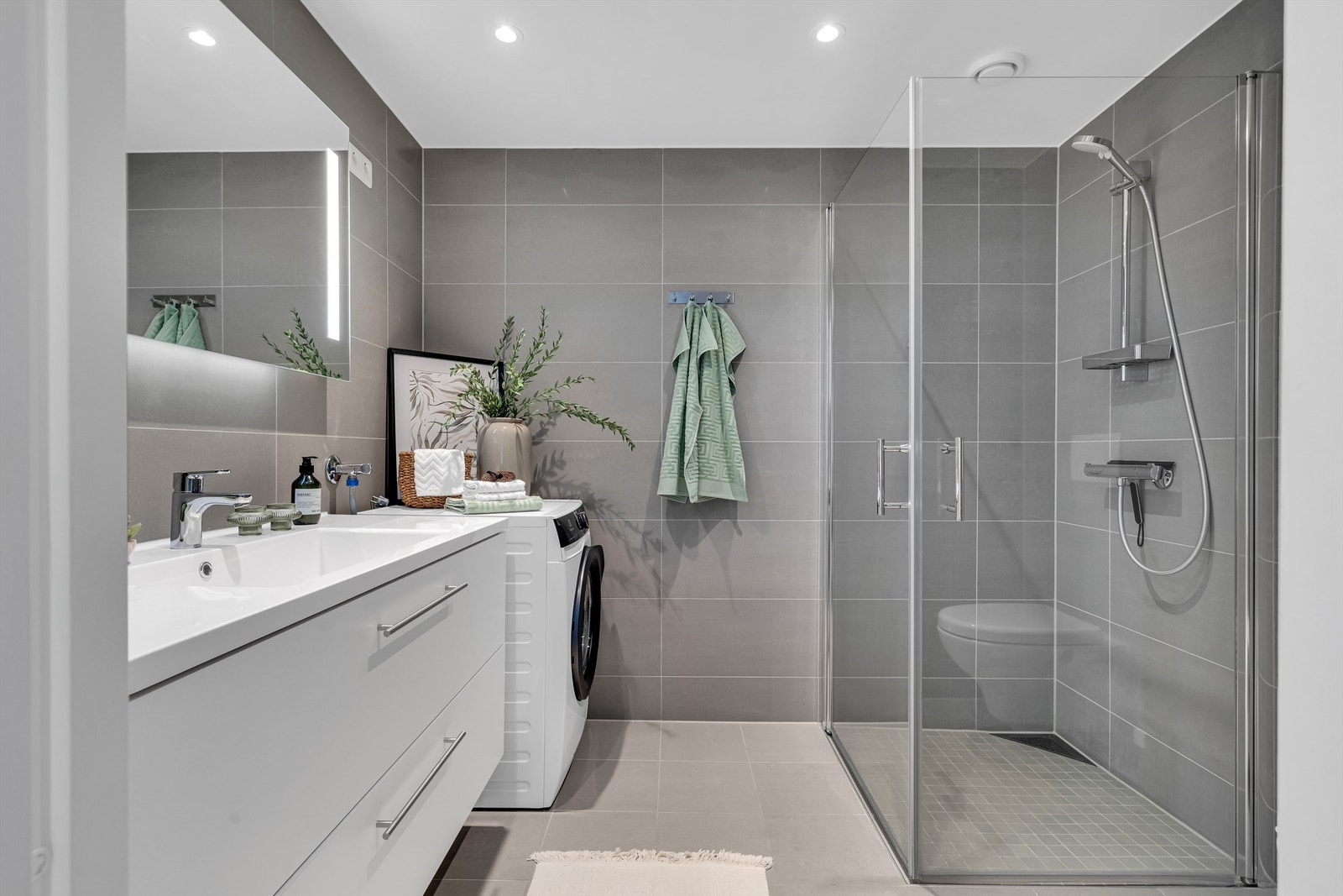 Innbydende og meget romslig bad med grå fliser på vegg- og gulvflater med varme i gulv og downlights i himling Galleribilde