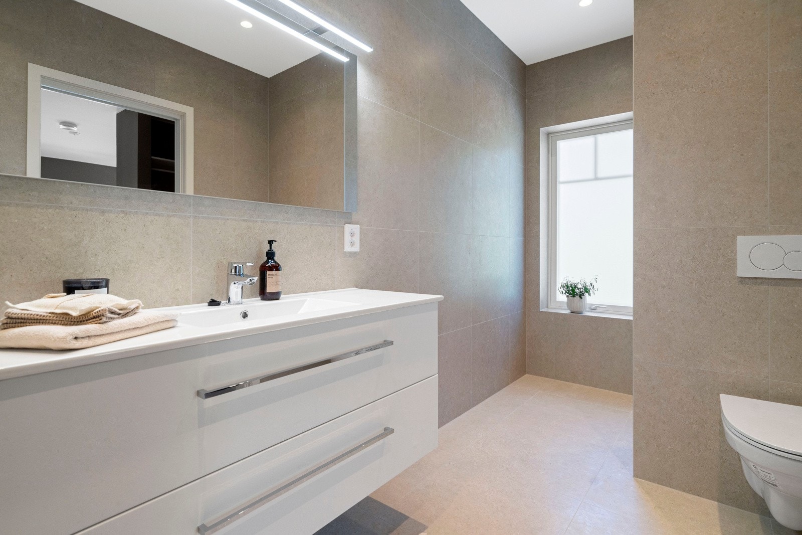 Begge badene er helfliset i moderne fargevalg og inneholder baderomsmøbler, dusj, vegghengt toalett og varmekabler i gulv Galleribilde