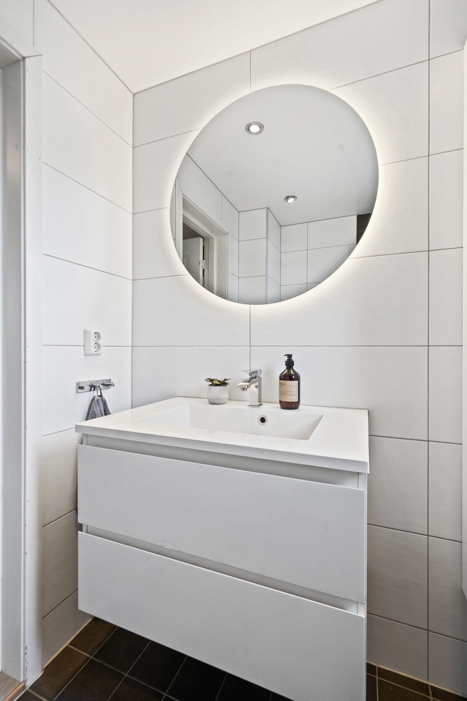 Innredningen består av servant med hvite skuffer, et høyskap og et rundt, stilig speil med bakenforliggende belysning Galleribilde