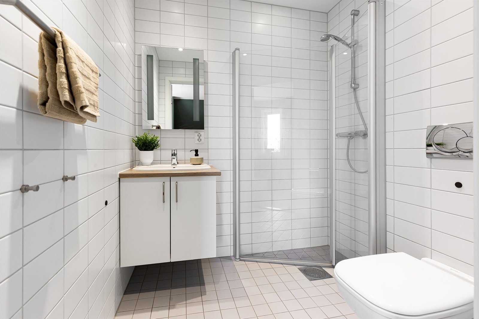 Baderom med dusjhjørne med dører i herdet glass. Benkeplate med nedfelt servant. Underskap. Speil med lys over servant. Vegghengt toalett. Galleribilde