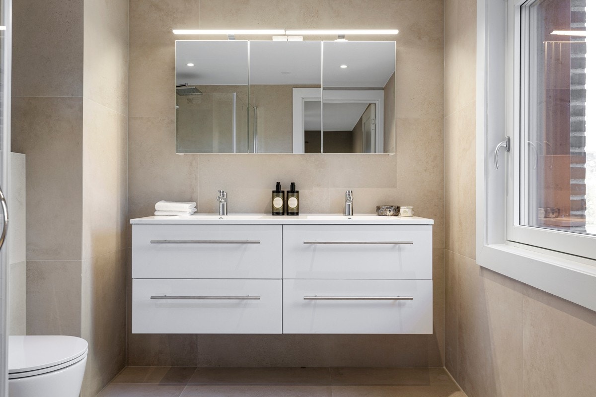 Videre har badet heldekkende dobbelservant på møblement med hvite skuffer. Over er det speilskap med belysning og tre dører Galleribilde