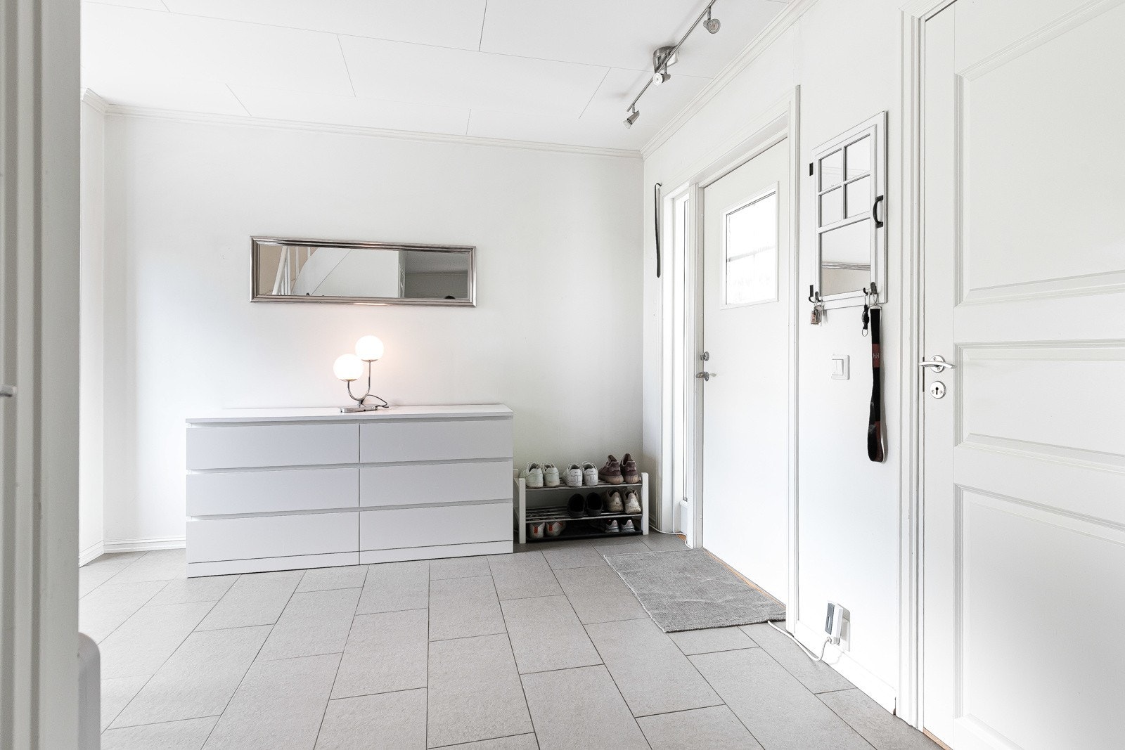 En ytterdør med sidevindu viser vei inn til en romslig entré, pent innredet med grå gulvfliser, gulvvarme og hvitmalte vegger. Galleribilde