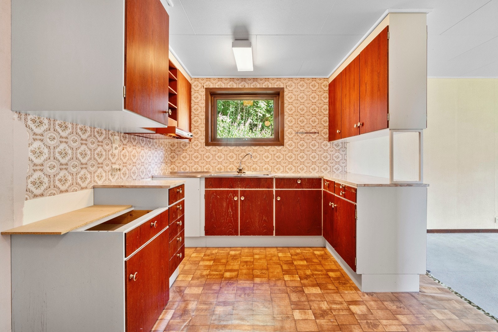 Eldre kjøkken innredet med under- og overskap samt benkeplate med oppvaskkum. Kjøkkenet har ingen hvitevarer. Galleribilde