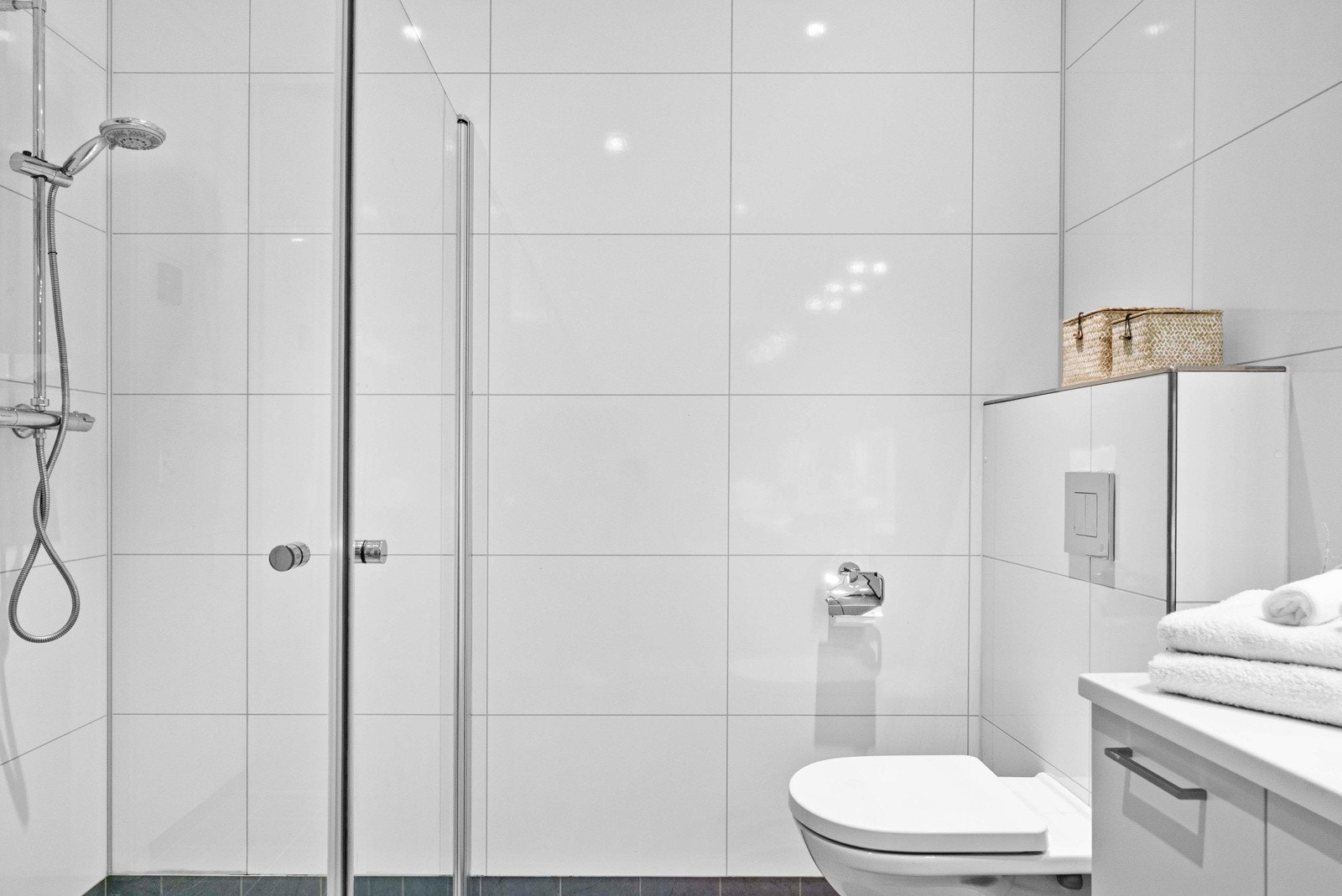 Innbydende bad med flislagt gulv og våtromspanel på veggene i lyse farger. Badet er ellers innredet med servant med underskap, speilskap, veggmontert dusj med dusjvegg og vegghengt toalett. Galleribilde