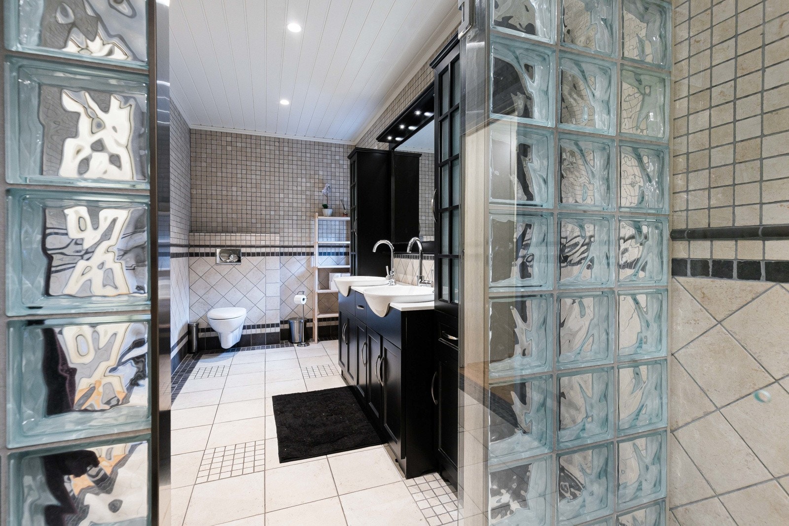 er det hvite fliser og mørke mosaikkfliser på gulvet. Veggene er kledd med fliser i sort og en nøytral sandtone. Galleribilde